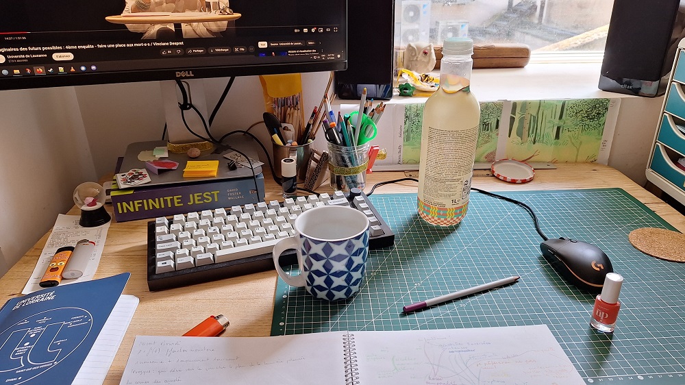 Une photographie de mon bureau, il y a le carnet aux feuilles blanches ouvert sur la carte heuristique, un flacon de vernis rose-orange, une tasse de café, un crayon violet, un briquet orange, le carnet univ-lorr, deux autres briques, l'escargot boule de neige offert par sarah, des trucs et des bidules encores fourmillent partout, sur l'écran de l'ordinateur un séminaire-conférence de Vinciane Desprets sur faire une place aux mortes