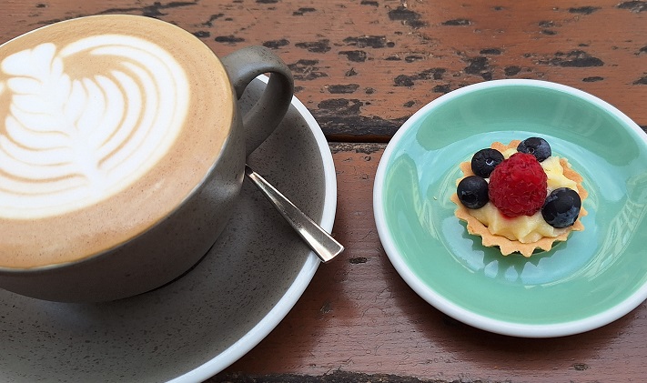 Une photographie d'une jolie tasse de café latte au lait d'avoine et une petite tartelette avec une frambroise et quelques myrtilles, la table en bois est un peu abîmé c'est joli