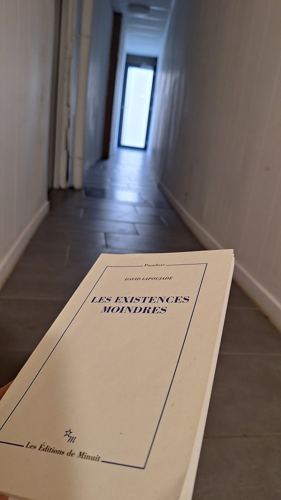 Une photo verticale dans le couloir du rez de chaussée de l'immeuble, avec en premier plan la couverture du livre Les existences moindres : c'est sombre et frais avec le carrelage gris et la lumière au bout du couloir vers l'extérieur