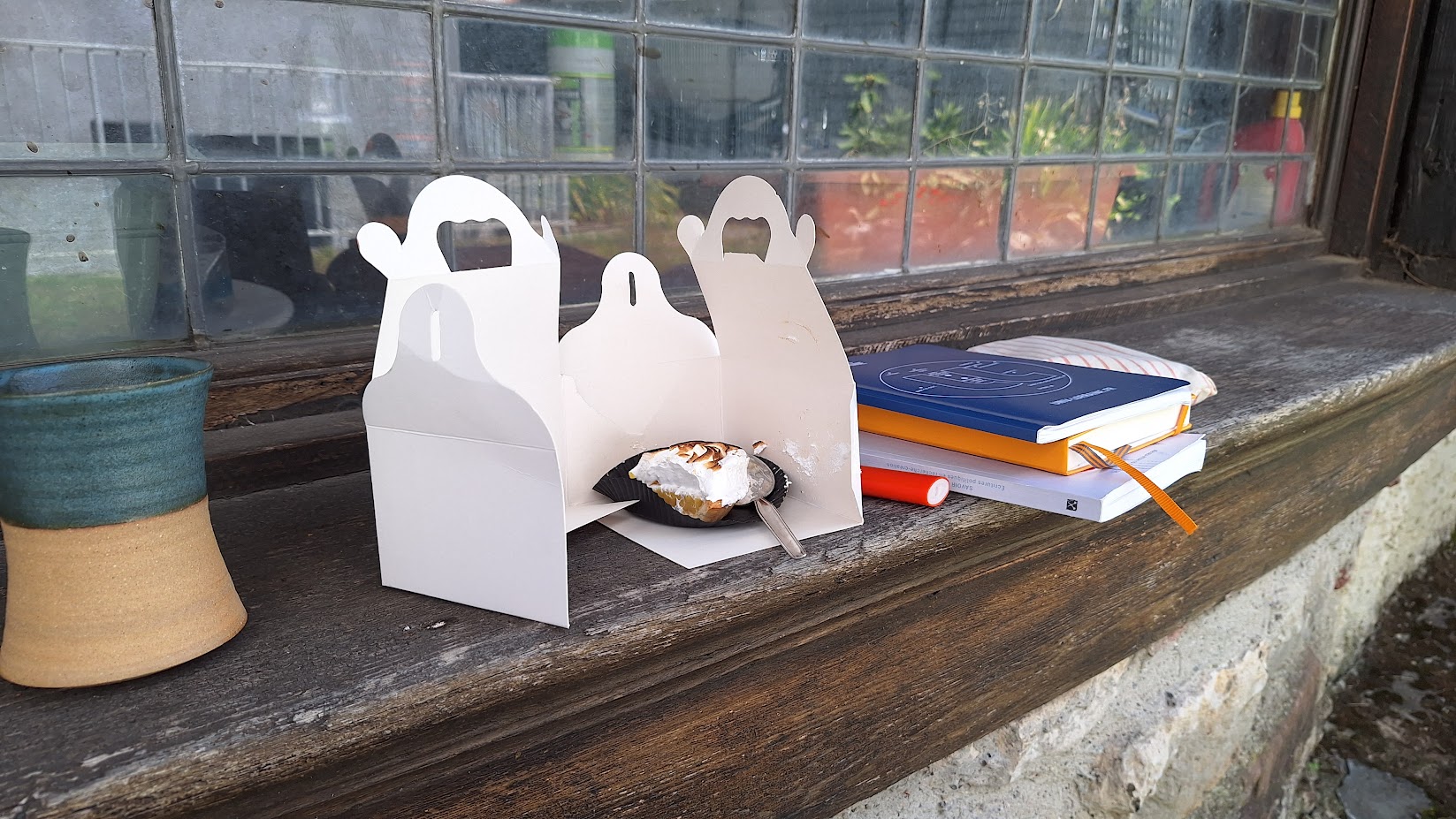 Une photographie d'un rebord de petit cabanon en bois, il y a dessus une tasse, une tarte au citron meringué entamé avec le carton déchiré autour, un briquet, un tas avec deux carnets et un livre, une trousse