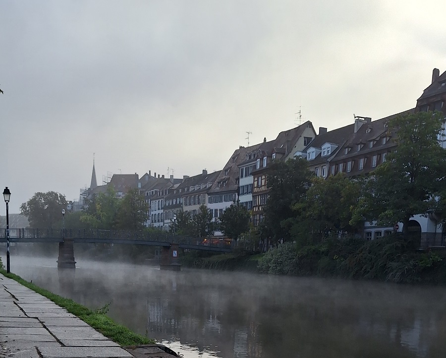 Une photographie prise dimanche matin à 8h38 le long de l'Ill à Strasbourg, il y a de la brume au dessus de la rivière, un petit pont au loin, le ciel est nuageux gris lumineux.