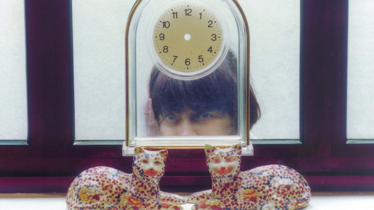 Image du film les Glâneurs d'Agnès Varda avec une horloge transparent sans aiguilles, un bout de visage d'Agnès Varda derrière et deux petites statuettes de félins devant